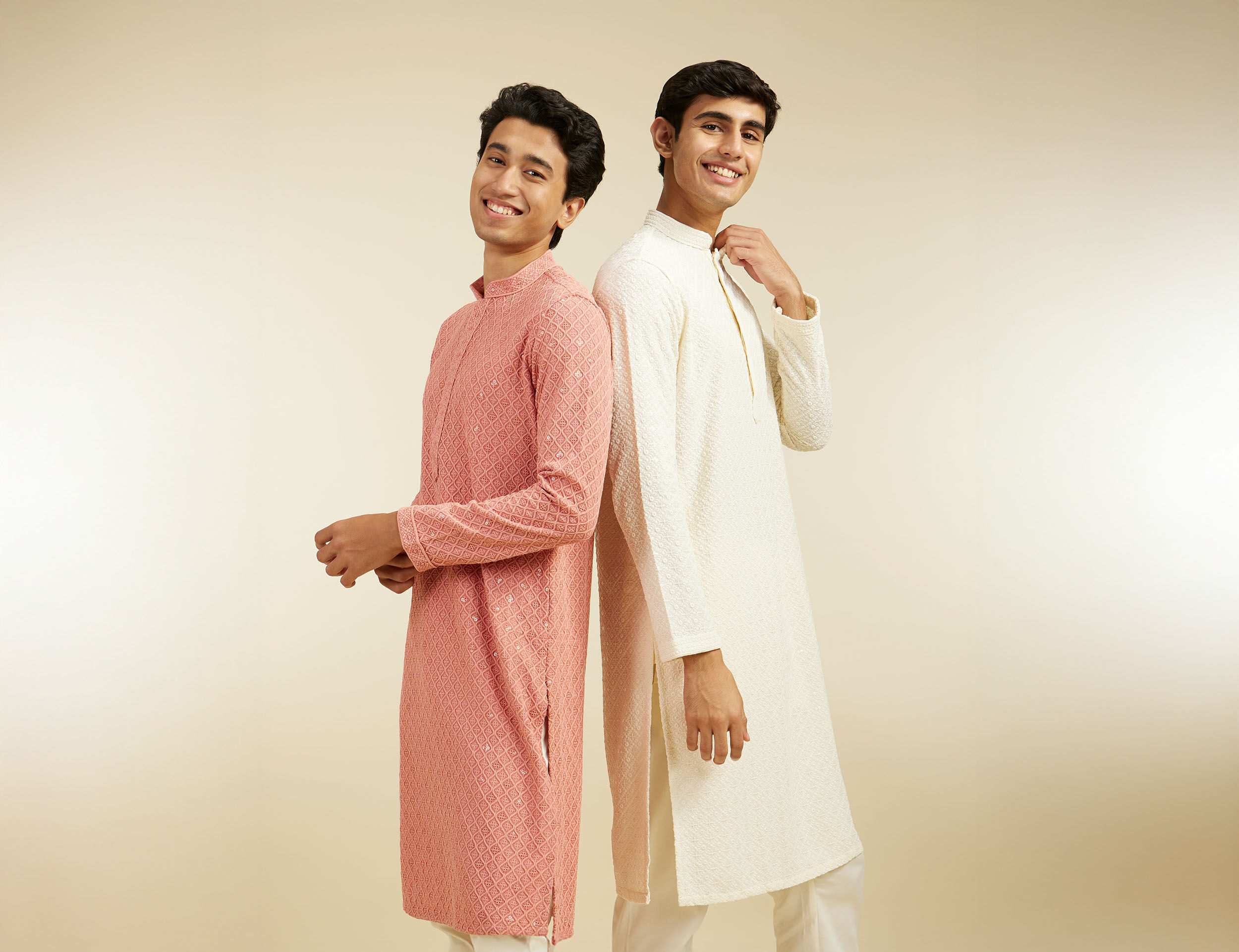 Two young men in pastel peach and off-white kurta pajamas smiling and posing back-to-back, showcasing elegant festive ethnic wear.